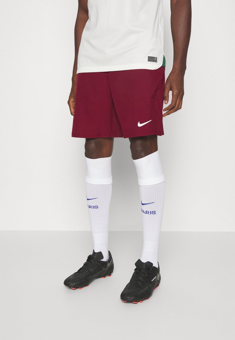 Personne portant un short Nike bordeaux, des chaussettes blanches montantes avec le texte « Paris » et des chaussures de football Nike noires, debout devant un fond uni.