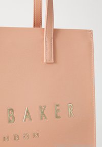 Návrh dámskej tašky v broskyňovej farbe z faux kože s dvojitými popruhmi, s embosovanou textúrou a zlatým metalickým nápisom "BAKER LONDON."