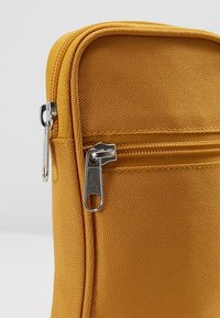 Gros plan d'une pochette en tissu jaune moutarde avec deux curseurs de fermeture éclair en métal argenté, un sur la poche avant et un sur le compartiment principal.