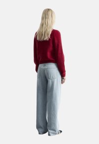 Roter, flauschiger Pullover mit weiten Ärmeln, kombiniert mit hellblauen weiten Jeans, von hinten gesehen, vor einem schlichten Hintergrund.