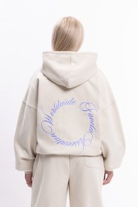 Béžová mikina s kapucňou vo voľnom strihu; na chrbte má fialový kruhový grafický nápis a malé ozdobné nitky pozdĺž dizajnu.