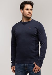 Granatowy sweter robiony na drutach z okrągłym dekoltem i mankietami ściągaczowymi. Posiada małe logo na piersi oraz dopasowaną sylwetkę. Noszony z niebieskimi dżinsami.