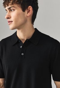 Polo noir en tissu tricoté, avec un col classique, une patte de boutonnage à trois boutons et des manches courtes avec des accents côtelés.