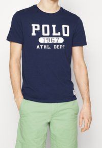 Navy blue cotton t-shirt featuring white printed text "POLO 1967 ATHL. DEPT." Round neck and short sleeves, paired with light green shorts.