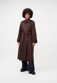 Cappotto trench in suede marrone con vita cintrata, ampio colletto e chiusura con bottoni doppi. Abbinato a jeans svasati scuri e stivali chunky.