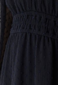 Marinblå klänning med textur, smockad midja och små broderade prickar. Har mjukt, krinklat tyg och en luftig silhuett.