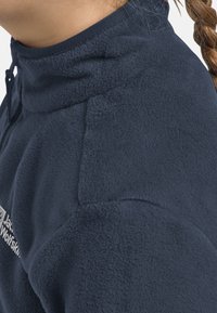 Giacca in pile blu navy con colletto alto, caratterizzata da una texture liscia e cuciture dettagliate. Include un logo sul lato anteriore.