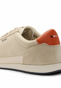Sneaker beige realizzata in materiali tessili e scamosciati, con un accentuato tallone arancione contrastante, suola in gomma nera e design minimale con branding.
