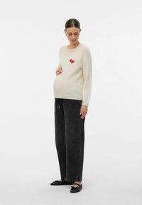 Mujer embarazada vistiendo un suéter crema con un corazón roto rojo, pantalones negros holgados y planos negros, de pie y sosteniendo suavemente su barriga.