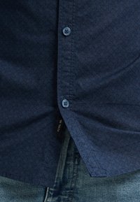 Dunkelblaues, knöpfbares Hemd mit geprägtem geometrischem Muster, ausgestattet mit kontrastierenden blauen Knöpfen, in der Nähe von Jeans im Denim-Stil mit sichtbaren Nähten.