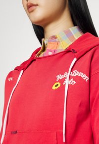 Červená mikina s bílými šňůrkami, s výšivkou „Ralph Lauren Polo“ a žlutou květinou. Nositelná přes vícebarevnou kostkovanou košili.