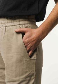 Beige broek met een gestructureerd ruitpatroon, uitgerust met een elastische tailleband en een zijzak. Een hand steekt in de zak.