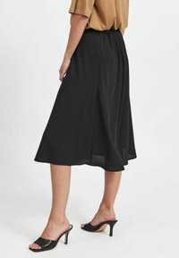 Jupe midi noire en tissu léger avec une taille élastique, présentant un design fluide et une silhouette simple, associée à des mules à talons noirs.