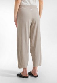 Nainen, joka seisoo valkoista taustaa vasten selin kuvattuna. Hänellä on päällään beige leveälahkeiset housut ja mustat slip-on kengät.