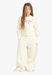 Ragazza giovane con capelli lunghi che indossa pantaloni larghi color crema e una felpa abbinata con logo viola, in piedi con le mani in tasca.