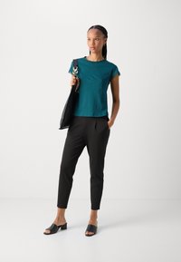 Teal Kurzarm-T-Shirt, kombiniert mit schwarzen schmal geschnittenen Hosen und schwarzen offenen Blockabsatz-Mules, gehalten mit einer schwarzen Schultertasche.