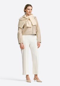 Veste de motard en cuir beige avec des détails de fermeture éclair, écharpe texturée crème, pantalon blanc ajusté et escarpins crème à bout pointu.
