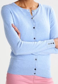 Ljusblå cardigan med knapprad, bärs av en person som korsar armarna, i kombination med rosa byxor mot en neutral bakgrund.