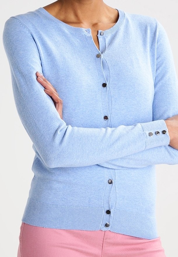 Ljusblå cardigan med knapprad, bärs av en person som korsar armarna, i kombination med rosa byxor mot en neutral bakgrund.