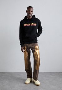 Sweatshirt à capuche noir avec un logo "glace" coloré, associé à un pantalon marron avec des panneaux latéraux métalliques dorés et des baskets beiges.