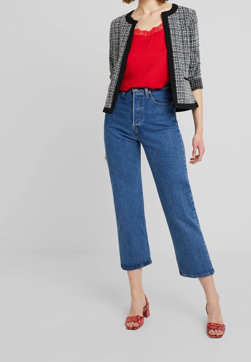 Femme portant une veste en tweed noir et blanc, un débardeur en dentelle rouge, un jean bleu taille haute et des talons ouverts rouges avec un motif serpent.