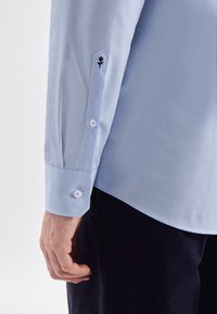 Camicia a righe azzurro chiaro con polsini abbottonati, caratterizzata da una forma esagonale e un dettaglio floreale ricamato sul polsino. Tessuto morbido.