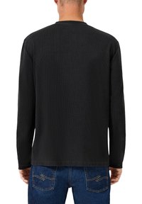Mies, who wears a plain black long-sleeve shirt and blue jeans with yellow stitching, seen from the back against a white background.