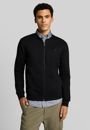 Junger Mann mit lockigem Haar, der eine schwarze Jacke mit Reißverschluss über einem grauen Hemd mit Knöpfen und beigen Cargohosen trägt, steht mit einer Hand in der Tasche.