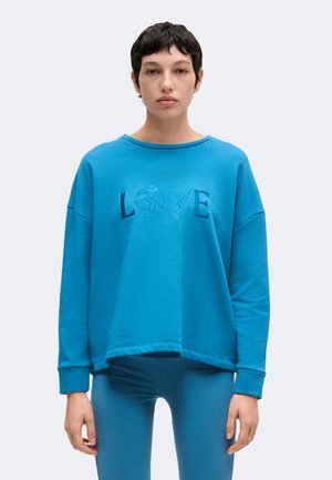 Person mit kurzen dunklen Haaren trägt ein weites blaues Sweatshirt mit der Aufschrift "LOVE" und passende blaue Leggings, steht vor einem schlichten Hintergrund.
