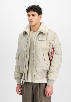 Beige bomber jacket with a fur-lined collar, zippered front, ribbed cuffs, and a pocket with a red tag detail. Smooth, shiny material.