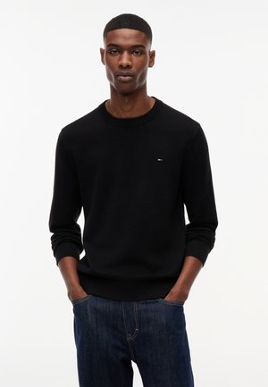 Junger Mann trägt einen schwarzen Langarmpullover mit kleinem Logo auf der Brust und blaue Jeans, Hände in den Taschen, schaut nach vorne.