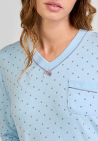 Top de color azul claro con escote en V y estampado de lunares grises, que presenta un pequeño lazo decorativo cerca del escote y un bolsillo en el pecho con detalle de ribete.