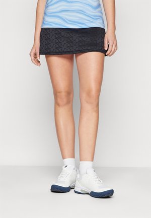 Femme portant une mini-jupe noire à motifs, un haut bleu, des chaussettes blanches et des chaussures de sport blanches avec des semelles foncées, debout devant un fond uni.
