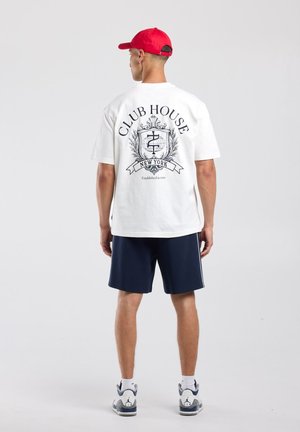 Mann trägt weißes Club House New York T-Shirt, marineblaue Shorts, rote Kappe und weiße Sneakers und steht mit dem Rücken zur Kamera auf einfarbigem Hintergrund.