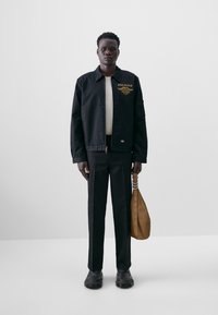 Black jacket with gold embroidery, fitted trousers, and black shoes. Brown bag with strap and eyelet details. Minimalist presentation.