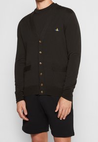 Svart stickad cardigan med v-ringning, fem bruna knappar, två framfickor och en liten broderad logotyp på vänster bröst.