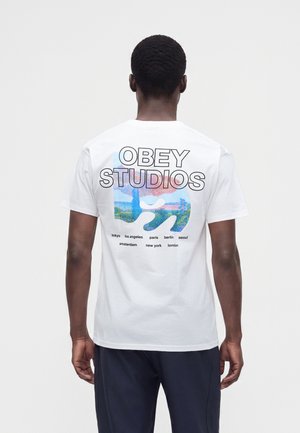 Persona que lleva una camiseta blanca con el texto "OBEY STUDIOS" y una gráfica colorida que muestra nombres de ciudades, incluyendo Tokio, Los Ángeles, París y Berlín, en la parte de atrás.