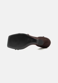 Zapato de tacón alto de ante marrón visto desde la suela, mostrando una parte inferior negra y lisa con marcas de tamaño y marca.