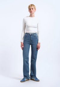 Weißes Langarmshirt, hochtaillierte blaue Jeans und blaue Sneaker mit grauen Streifen. Glatter Stoff, gerade Beinschnitt, minimalistische Hardware.