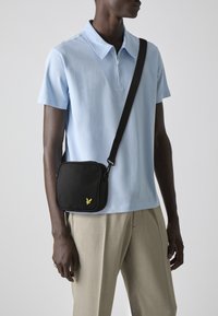 Zwarte schoudertas met rits, gele logo-details, zachte stof, rechthoekige vorm, gedragen over een lichtblauwe polo en beige broek.