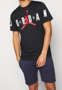 Czarna bawełniana koszulka z dużym białym napisem "JORDAN" i czerwoną grafiką jumpmana. Noszona z ciemnoniebieskimi szortami, gładka tekstura.