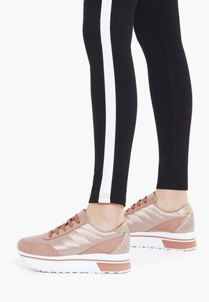 Chaussures de sport rose clair avec une finition texturée, dotées d'une semelle blanche et d'un petit talon, associées à un legging noir avec des rayures blanches.