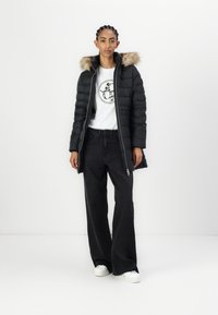 Žena u crnoj jorkerici s krznenom kapuljačom, bijeloj grafičkoj majici, širokim crnim trapericama i bijelim tenisicama, stoji pred bijelom pozadinom.