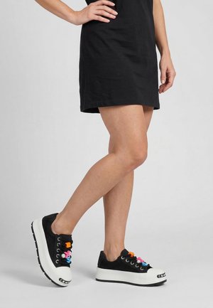 Persoon die een korte zwarte jurk draagt en zwarte platformsneakers met kleurrijke letterbedeltjes aan de veters, staand tegen een effen achtergrond.