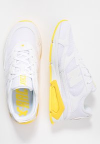 Des chaussures de sport blanches avec des accents jaunes, une tige en mesh, une semelle texturée et un logo "N" bien en vue sur le côté. Lacets et col rembourré inclus.