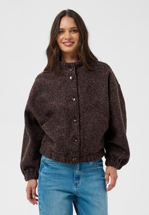Femme aux longs cheveux foncés portant une veste en laine marron foncé boutonnée et un jean bleu, souriante sur un fond clair uni.