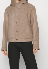 Man som bär en ljusbrun knäppbar ulljacka med huva och svarta byxor, stående mot en neutral bakgrund.