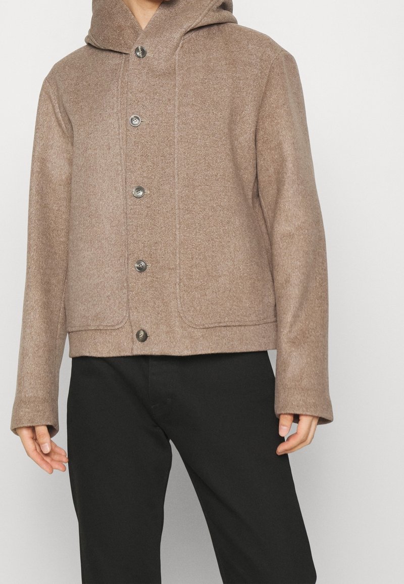 Man som bär en ljusbrun knäppbar ulljacka med huva och svarta byxor, stående mot en neutral bakgrund.