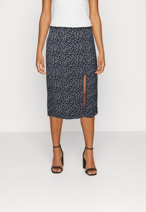 Woman wearing a black midi skirt with a white floral pattern and front slit, paired with black high-heeled sandals and a white sleeveless top.