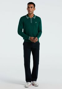 Grünes Langarm-Polohemd mit Kragen und weißen Akzenten, kombiniert mit dunklen Hosen und weißen Sneakers. Das Material wirkt glatt und gestrickt.